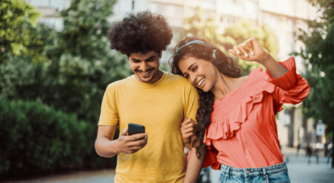 Two young people walking outdoors, smiling and looking at a smartphone together. One wears a yellow t-shirt, the other a coral ruffled top with headphones on.