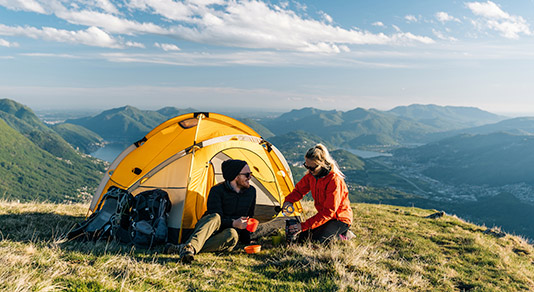 Couple camping on mountain top, prepare food and beverages next to tent.