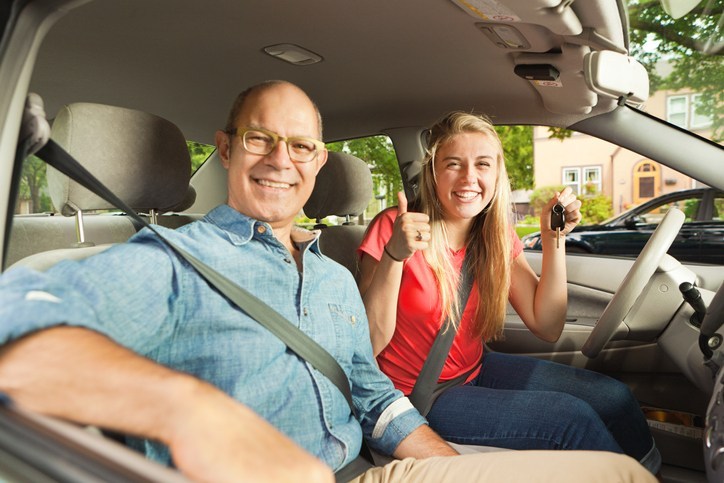 A happy teenager young woman receiving a new car from her parent father. The young woman is holding the key to the car and they are smiling and posing for the camera inside their new car.