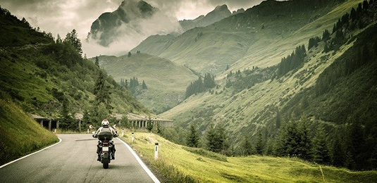 Man riding a motor cycle on a raod between lush green mountains